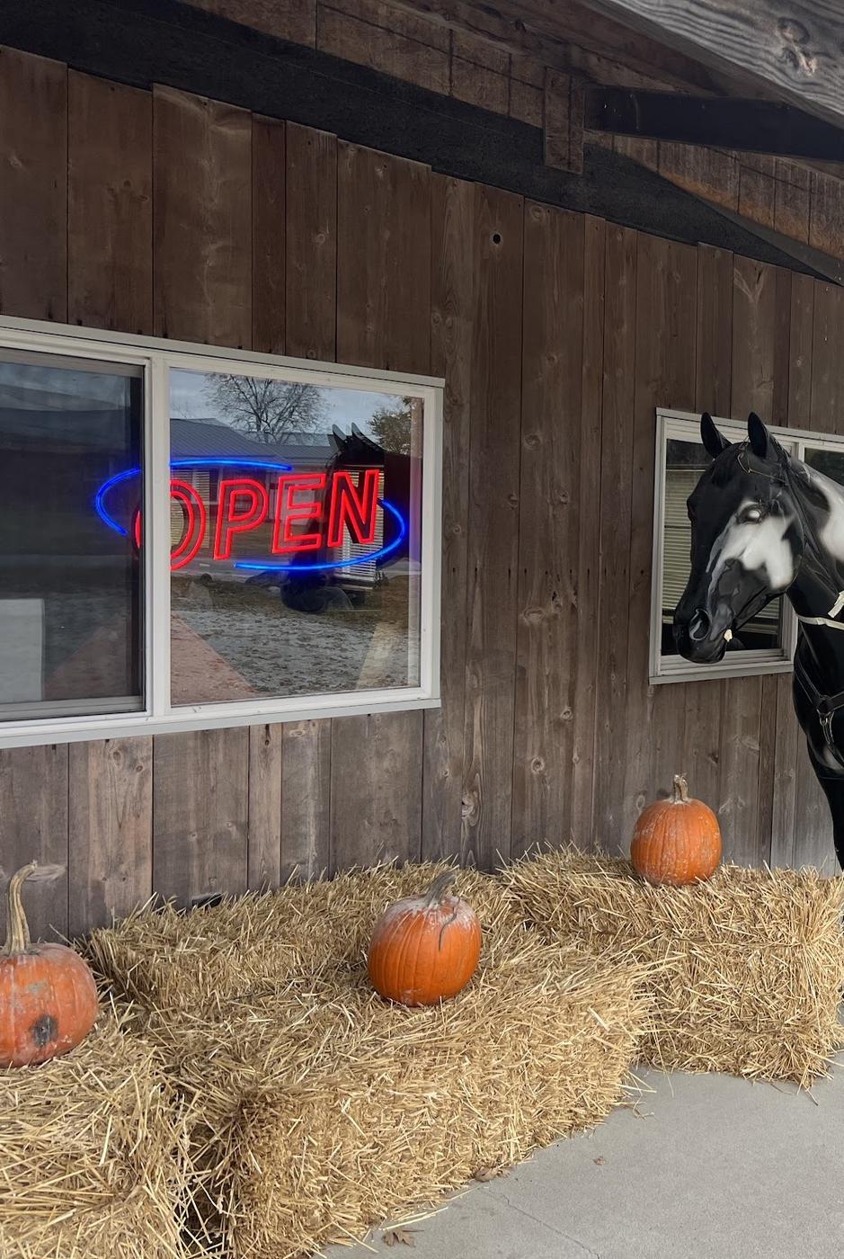 The front of the building: dark barn-wood siding, a covered porch, a neon open sign in the window, straw bales along the wall, and the head of the life-size black and white horse that stands out front.