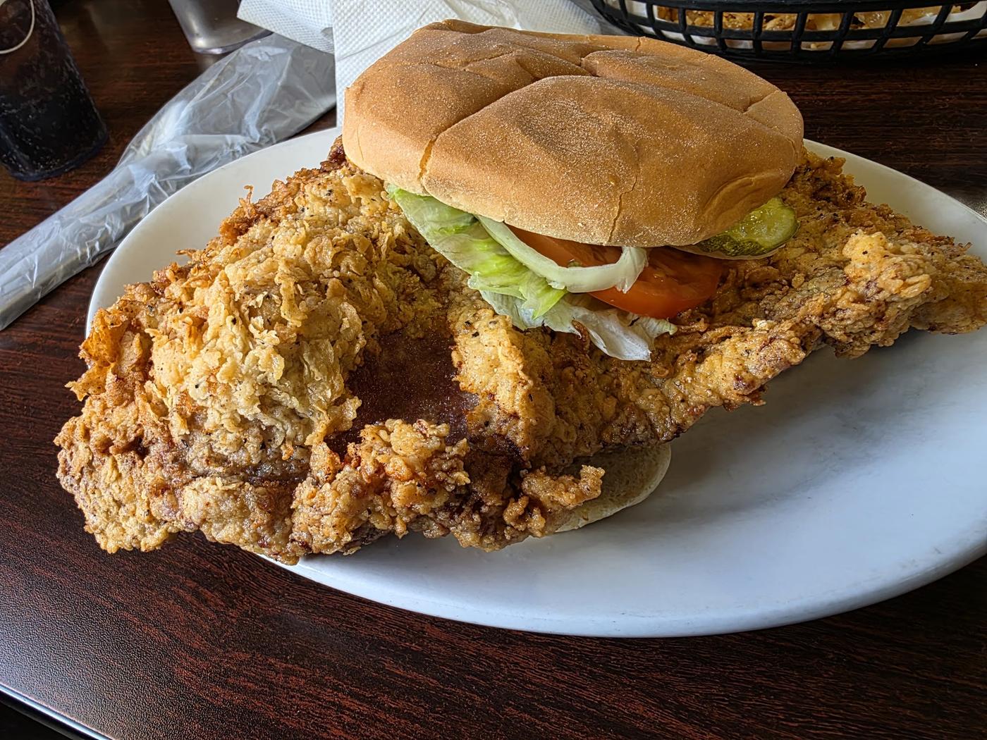 A hand-breaded tenderloin sandwich: a large craggy breaded cutlet overhanging a small bun on every side, with lettuce, tomato and a pickle, on a white plate.