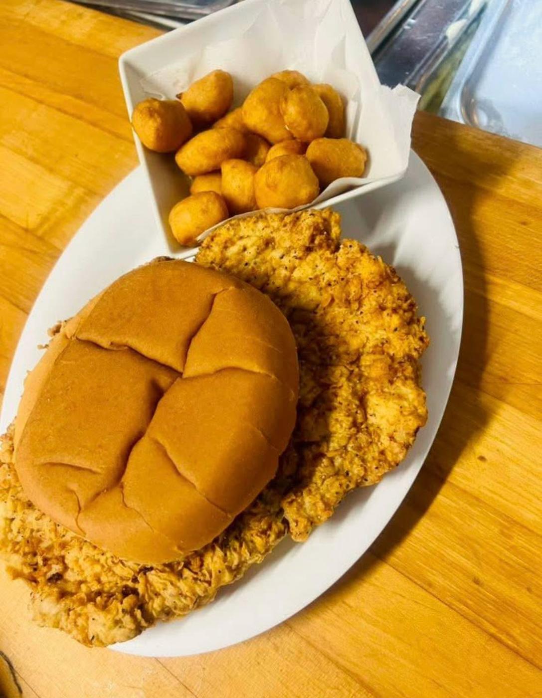 A hand-breaded tenderloin on butcher block, overhanging its bun, with a square dish of golden battered nuggets alongside.